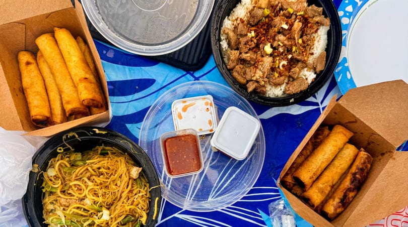 An overhead shot of a takeout meal with spring rolls, noodles, beef and rice, and sauces.