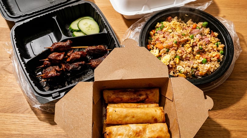 Takeout containers filled with fried rice, spring rolls, and meat skewers with cucumber slices.