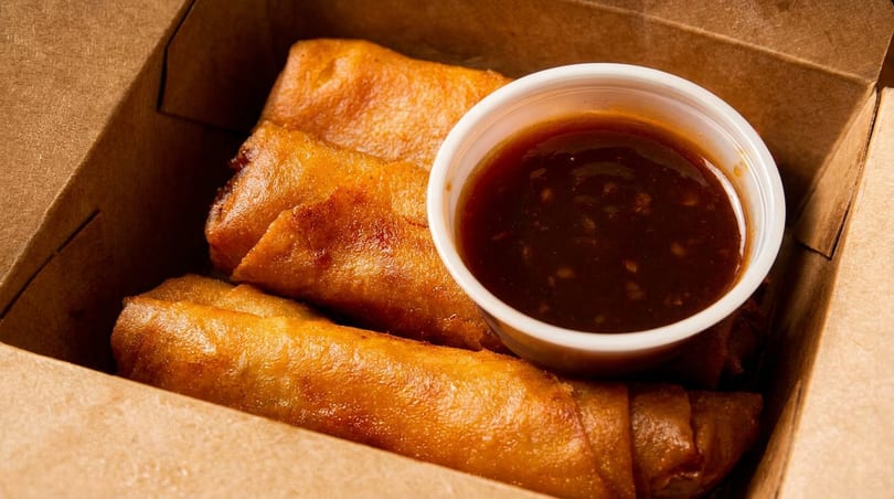 Golden-brown spring rolls and a small cup of dipping sauce in a brown takeout box.